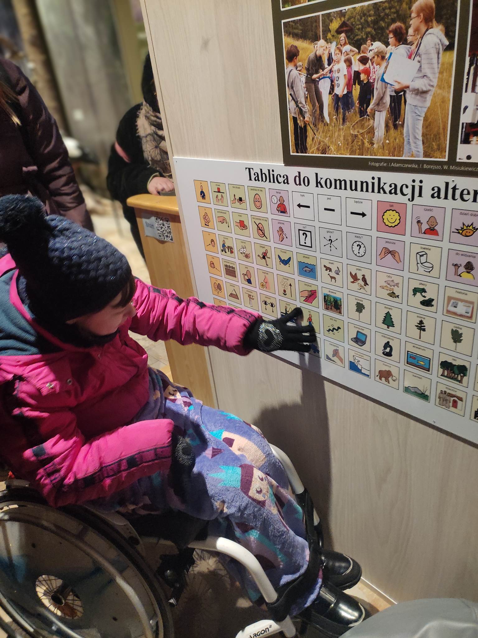 Kobieta na wózku inwalidzkim wskazuje na jeden z obrazków na tablicy do komunikacji alternatywnej.