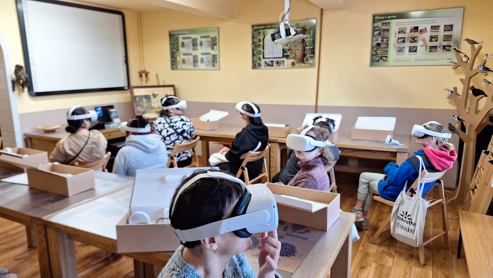Grupa osób siedząca w sali edukacyjnej ma na głowie okulary Virtual Reality. W sali na ścianach tablica interaktywna, plansze edukacyjne ze zwierzętami.