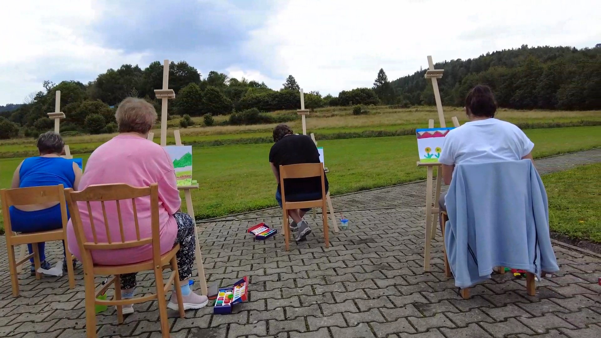 Cztery kobiety odwrócone tyłem siedzą na krzesłach i malują obrazy na drewnianych sztalugach. W tle krajobraz górski.