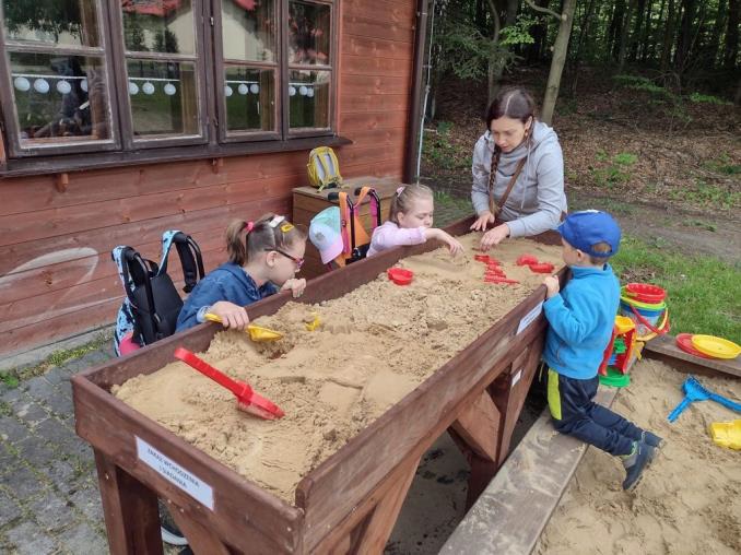 Piaskownica z nadstawką przy ścieżce sensorycznej. Drewniana piaskownica z nadstawką, przy której bawią się dzieci siedzące na wózkach.