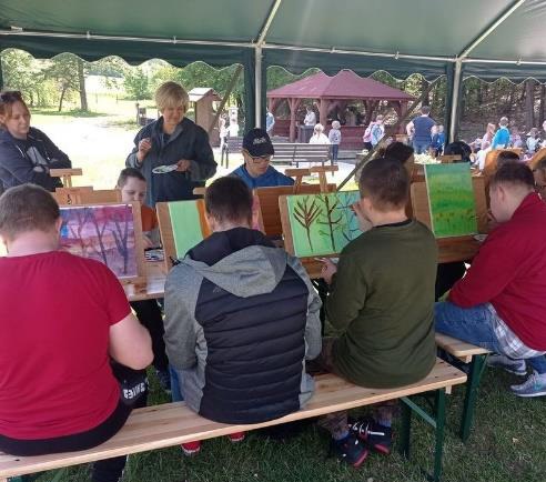 Plener malarski. Grupa osób maluje obrazy na drewnianych sztalugach pod zielonym namiotem.