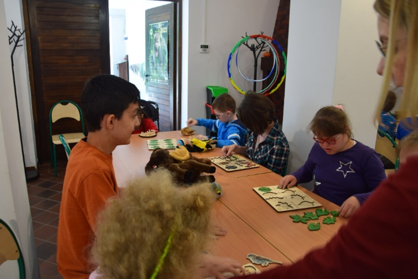 Sala edukacyjna, w której grupa dzieci wraz z opiekunem siedzi przy stole i uczestniczy w zajęciach edukacyjnych. W tle otwarte drewniane drzwi.