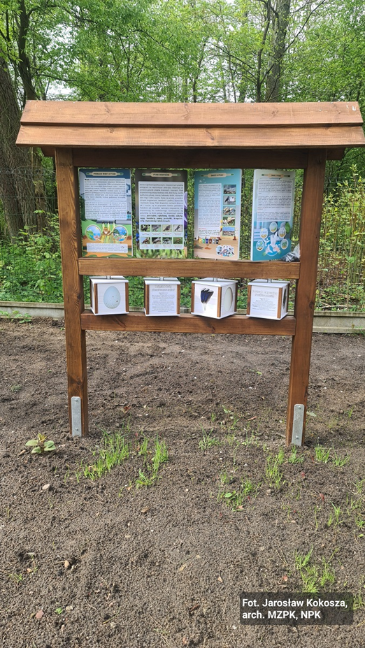 Drewniana tablica edukacyjna stojąca wśród drzew. W górnej części obracalne panele z opisami, w dolnej obracalne kostki z rysunkami. 