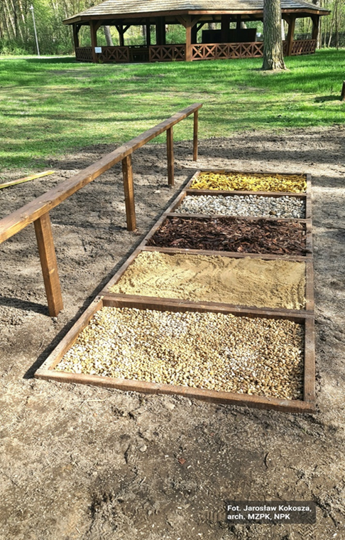 Ścieżka sensoryczna z różnymi rodzajami podłoża: żwir, piasek itp. Obok niej drewniana barierka. W tle na polanie leśnej duża drewniana wiata. 