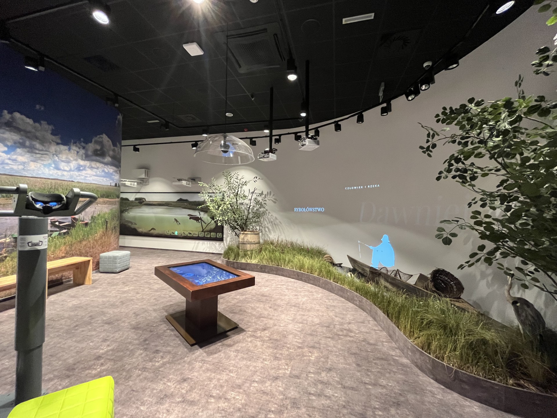 Wystawa stała dostępna dla osób z niepełnosprawnościami w Ośrodku Edukacji Przyrodniczej „Młynarzówka”. Fragment wystawy w budynku Ośrodka Edukacji Przyrodniczej "Młynarzówka" w Narwiańskim PN. Na środku tablica multimedialna na postumencie. W tle krajobraz rzeki z eksponatami łodzi, beczki i roślinnością nadrzeczną. Z lewej strony luneta do oglądania krajobrazu.