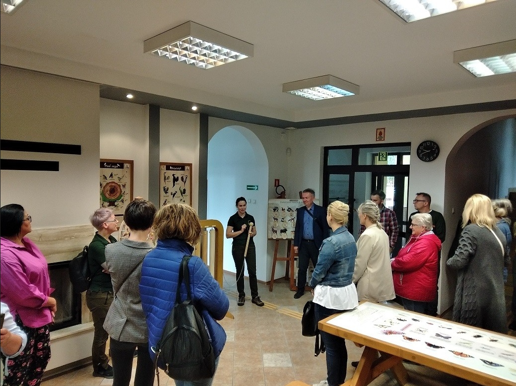 Fragment wystawy z grupą zwiedzających w budynku Ośrodka Edukacji Przyrodniczej w Pszczewie. Na środku przewodniczka mówiąca do grupy i pokazująca eksponat. Na ścianach tablice edukacyjne. Po prawej stronie drewniany stół z grafikami motyli.
