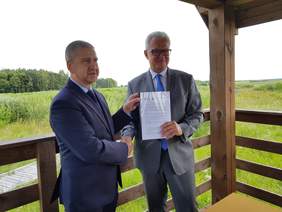 Pokaż zdjęcie: 2 mężczyzn w garniturach uściskujących sobie dłonie w geście gratulacji. Wspólnie prezentują trzymany przez siebie dokument. W tle zielony plener.