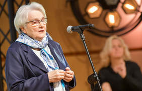 Pokaż zdjęcie: Kobieta o siwych włosach, przed nią mikrofon. 