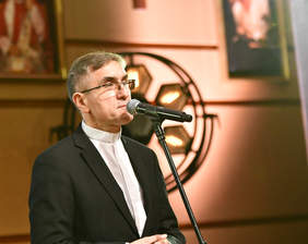 Pokaż zdjęcie: Mężczyzna w koszuli z koloratką, przed nim mikrofon