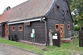 Pokaż zdjęcie: Drewniany budynek z czerwonym dachem. Na ścianie zamontowane urządzenie AED w zielonej zabudowie. Przed budynkiem znajduje się ławka oraz brukowany chodnik.