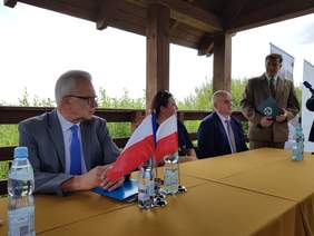 Pokaż zdjęcie: Przemawiający przed zgromadzonymi przy stole z flagami Unii Europejskiej i Polski, mężczyzna w służbowym garniturze. W dłoniach trzyma teczkę z opisem – Poleski Park Narodowy. Za nim 2 duże bannery i plenerowe pejzaże.