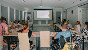 Pokaż zdjęcie: Grupa osób siedzi przy stole w sali patrząc na wyświetlany slajd.