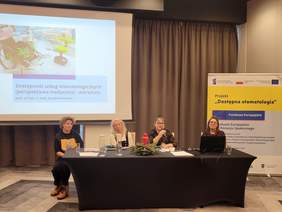 Pokaż zdjęcie: W auli przy stole prezydialnym siedzą 4 osoby. Jedna z nich mówi do mikrofonu. W tle ekran z prezentacją.