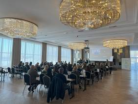 Pokaż zdjęcie: Ludzie siedzący tyłem na sali konferencyjnej słuchają znajdujących się na podwyższeniu prelegentów. Obok prelegentów biały ekran i roll-up. Na suficie kryształowe żyrandole. W tle okna z zasłonami. 