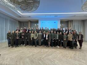 Pokaż zdjęcie: grupa ok. 50 osób ubranych głównie w zielone mundury lub polary z emblematami parków. Stoją na podwyższeniu i parkiecie przed nim. W tle biały ekran z napisem "OBSZAR CHRONIONY OBSZAR DOSTĘPNY. Na suficie kryształowy żyrandol. Po bokach okna 