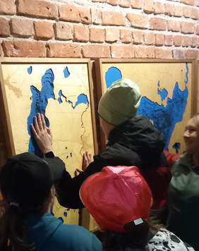 Pokaż zdjęcie: Grupa dzieci dotyka zawieszonej na ścianie mapy przedstawiającej zarys lądów i zbiorników wodnych.